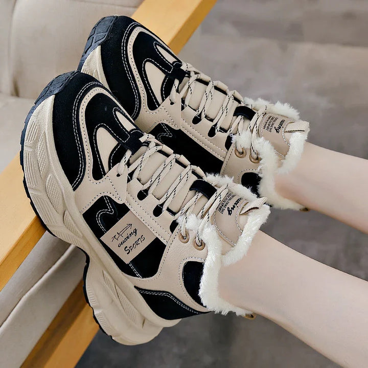 Beige and black fur-lined sports sneakers with thick soles worn on feet near wooden furniture