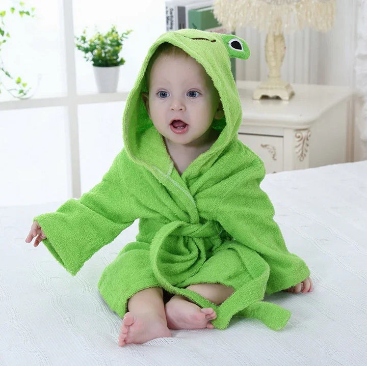 Baby sitting on bed wearing a green hooded frog bathrobe with eyes and mouth design indoors