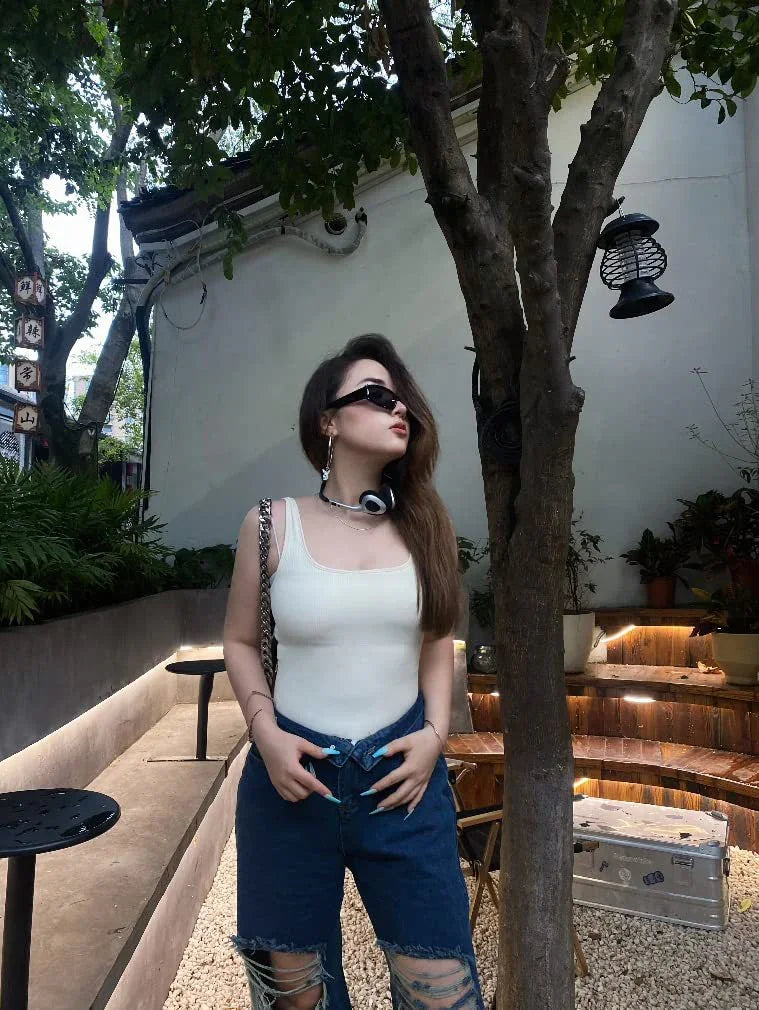 Young woman in white tank top and ripped jeans posing outdoors near tree and wooden seating