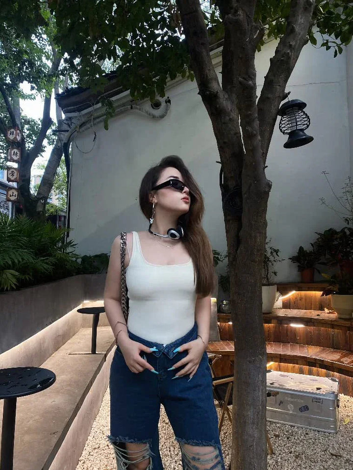 Young woman in white tank top and ripped jeans posing outdoors near tree and wooden seating