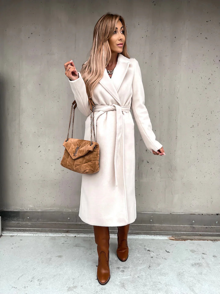Woman in white belted coat with tan boots and quilted brown handbag standing against gray concrete wall
