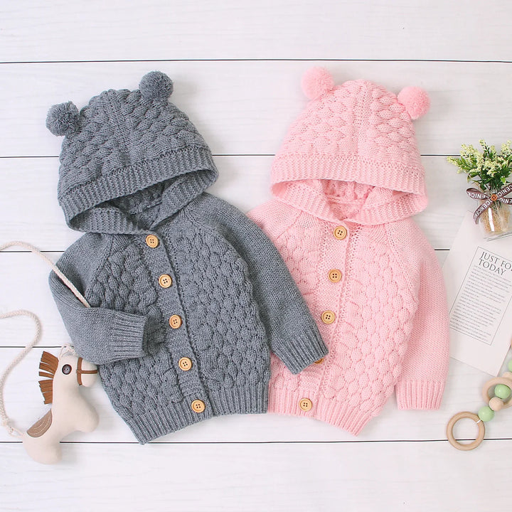 Gray and pink knitted baby cardigans with hoods and pom-poms on white wooden background