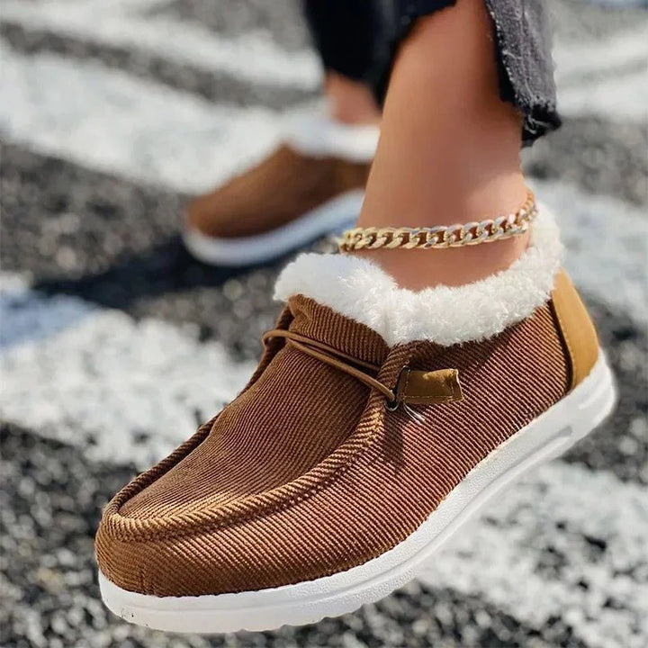 Close-up of a foot wearing brown textured slip-on shoes with white fur lining and a gold chain anklet