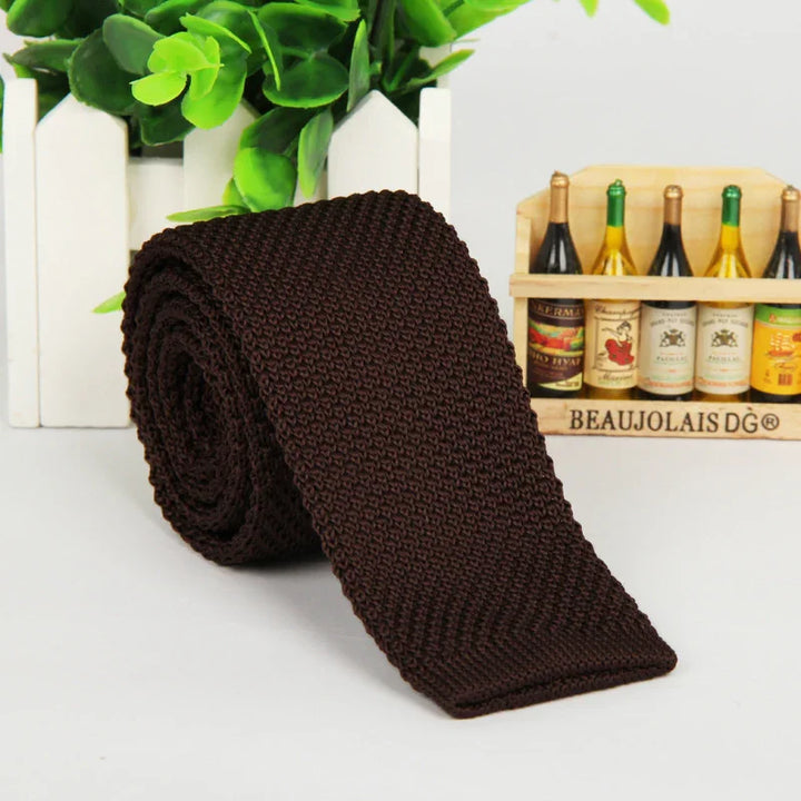 Dark brown knitted men's tie rolled up with green plant and miniature wine bottles background