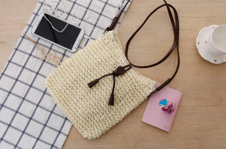 Woven beige crossbody bag with brown strap on wooden table with phone, pink notebook, and checkered cloth