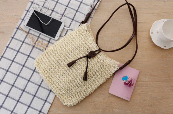 Woven beige crossbody bag with brown strap on wooden table with phone, pink notebook, and checkered cloth