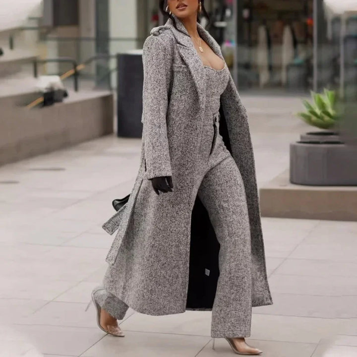 Woman walking in urban setting wearing long gray coat, matching pants, black gloves, and transparent high heels