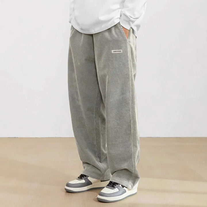 Person wearing loose gray corduroy pants with a small patch and gray-and-white sneakers on a beige floor