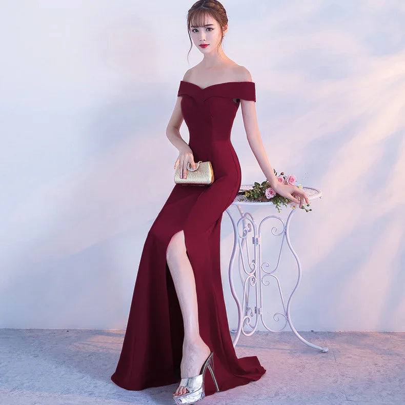 Woman in elegant off-shoulder burgundy evening gown with thigh slit, holding gold clutch and wearing silver heels