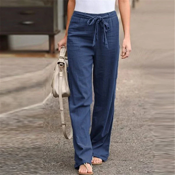 Woman wearing blue drawstring linen pants, white top, sandals, and holding beige handbag on street