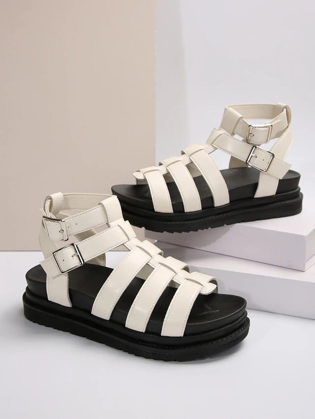 White strappy gladiator sandals with black cushioned soles and silver buckles on white background
