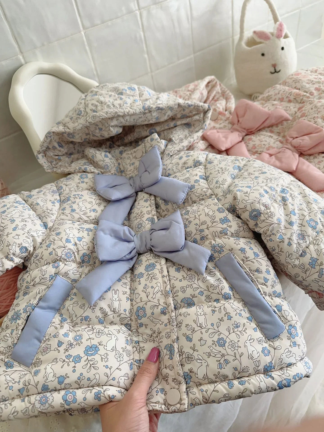 Hand holding a padded baby jacket with blue floral and bunny print, featuring blue bows and a hood, white tiled background