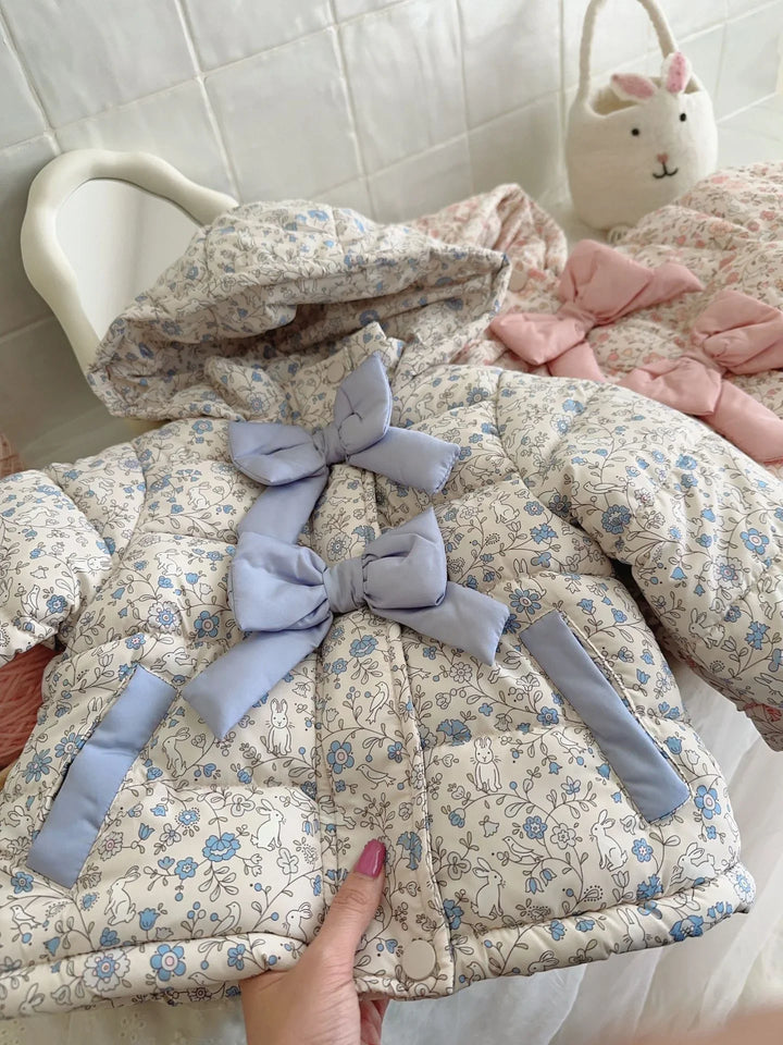 Hand holding a padded baby jacket with blue floral and bunny print, featuring blue bows and a hood, white tiled background