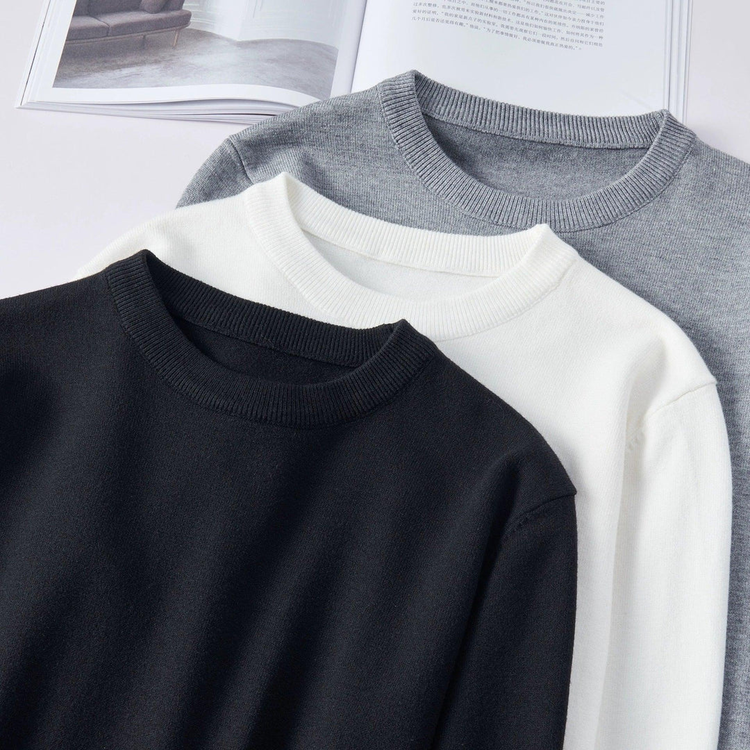 Three folded crewneck sweaters in black, white, and gray on a white surface with open book