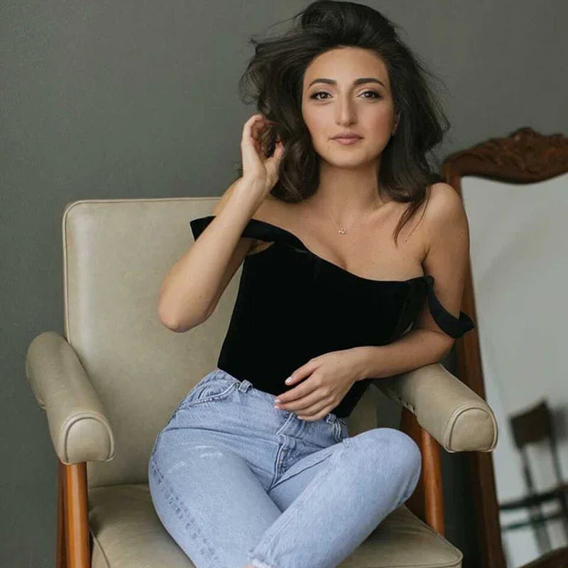 Young woman with dark hair in black off-shoulder top and light blue jeans sitting on beige armchair