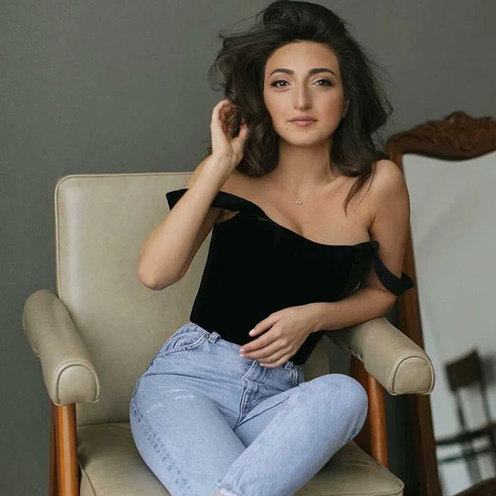 Young woman with dark hair in black off-shoulder top and light blue jeans sitting on beige armchair