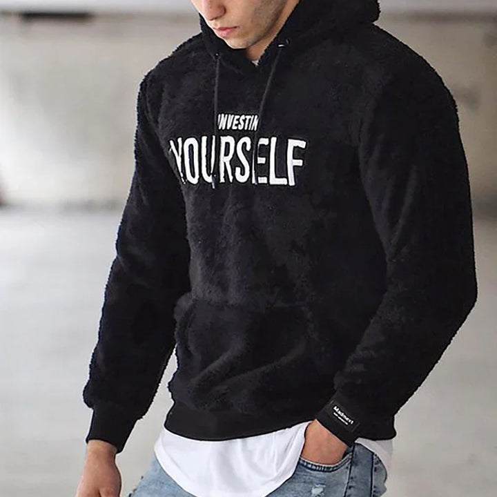 Man wearing black fuzzy hoodie with white text 'INVESTIN YOURSELF' in casual indoor setting