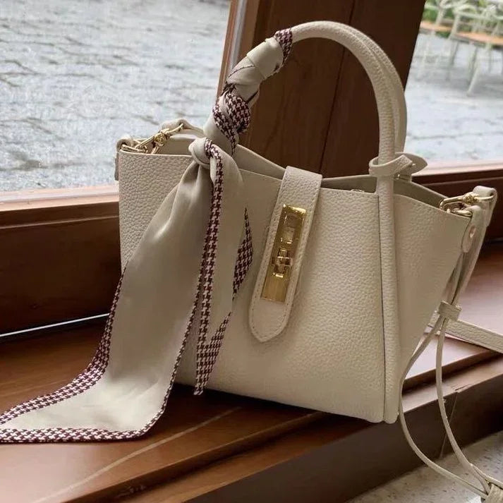 Elegant cream leather handbag with gold clasp and patterned silk scarf on wooden windowsill