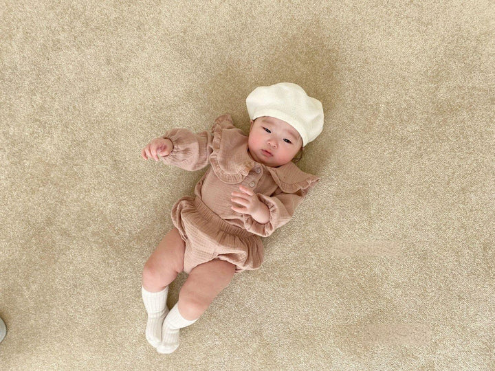 Baby in beige outfit and white beret lying on light brown carpet