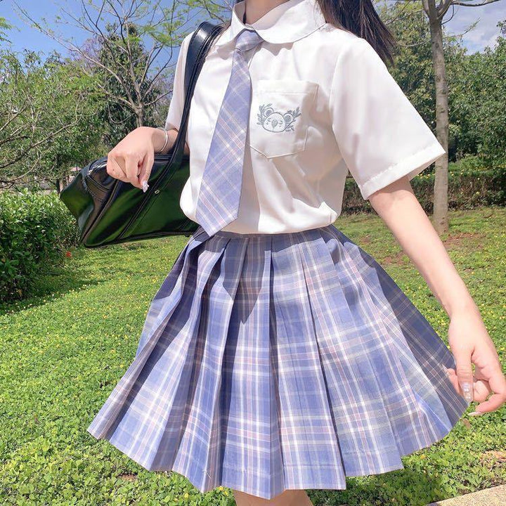 Young woman wearing a plaid blue pleated skirt and matching tie with a white blouse, outdoors with green grass and trees