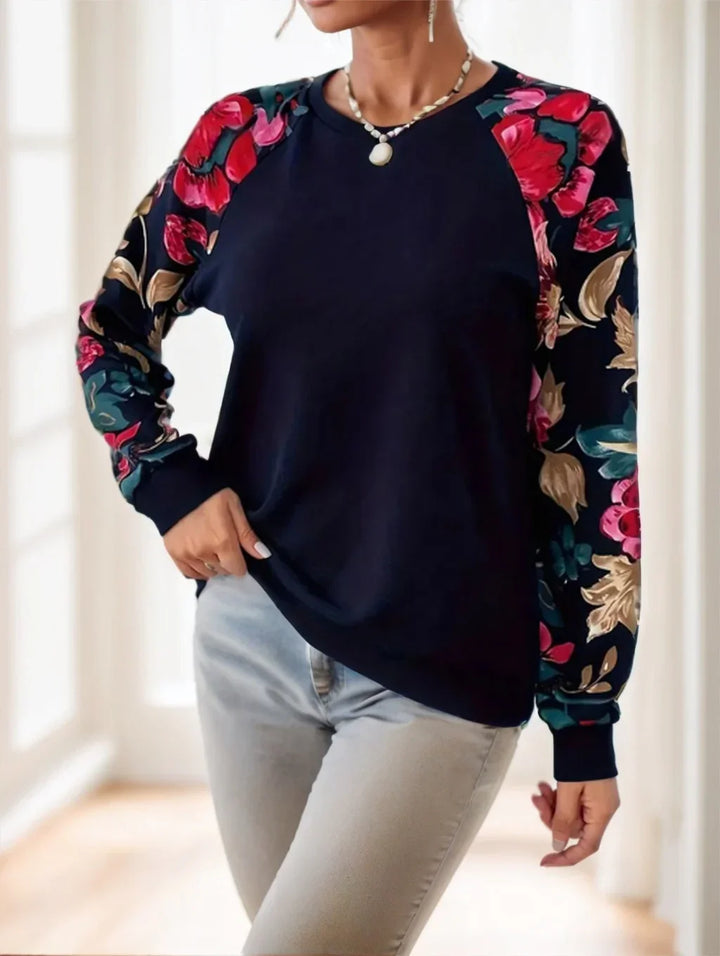 Woman wearing a navy blue sweatshirt with vibrant floral print sleeves and light blue jeans in bright indoor setting