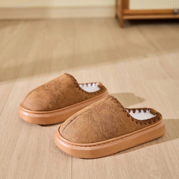 Pair of brown suede indoor slippers with thick soles and white fleece lining on wooden floor