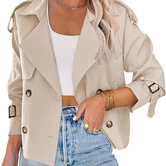 Woman wearing beige cropped trench coat with buckled cuffs, white crop top, and high-waisted blue jeans