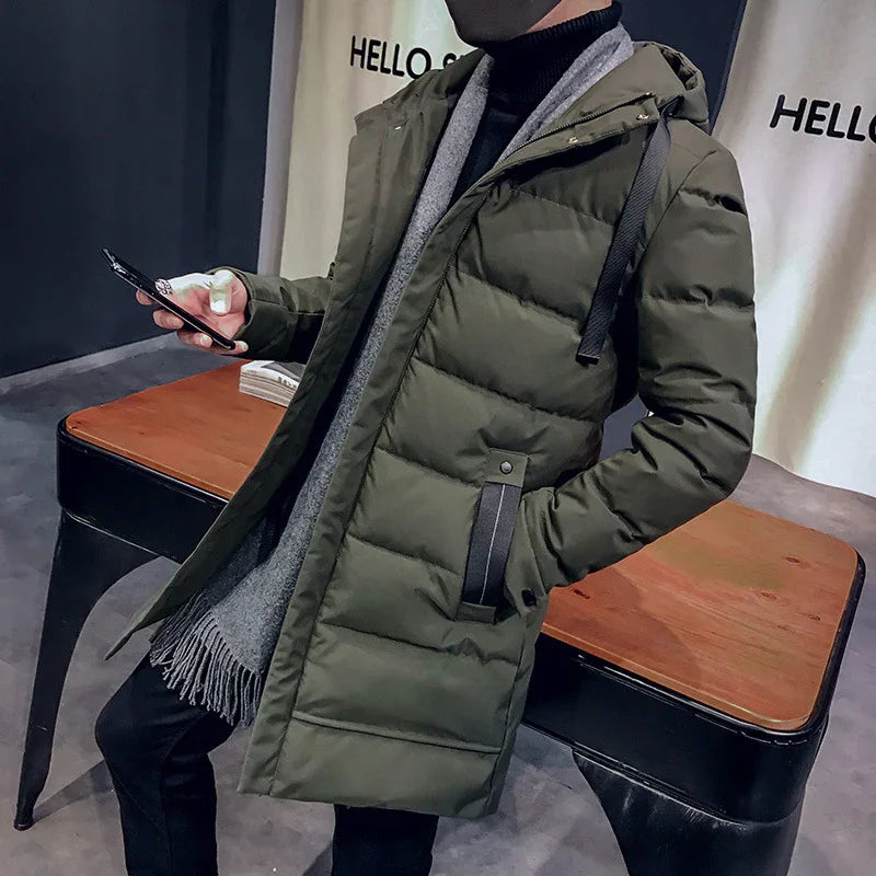 Man wearing olive green hooded puffer coat with gray scarf, using smartphone indoors by wooden table