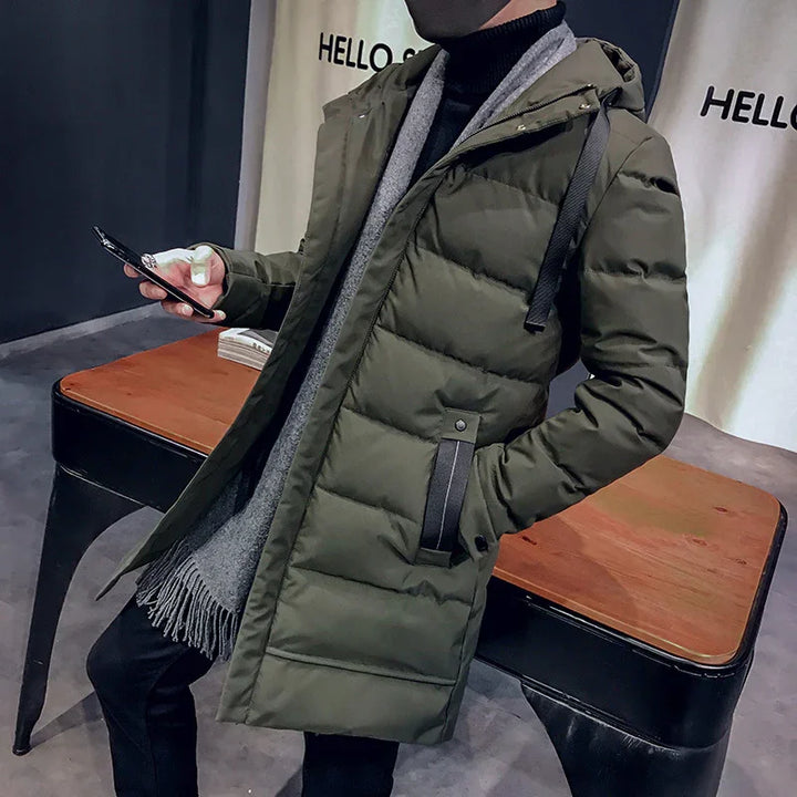 Man wearing olive green hooded puffer coat with gray scarf, using smartphone indoors by wooden table
