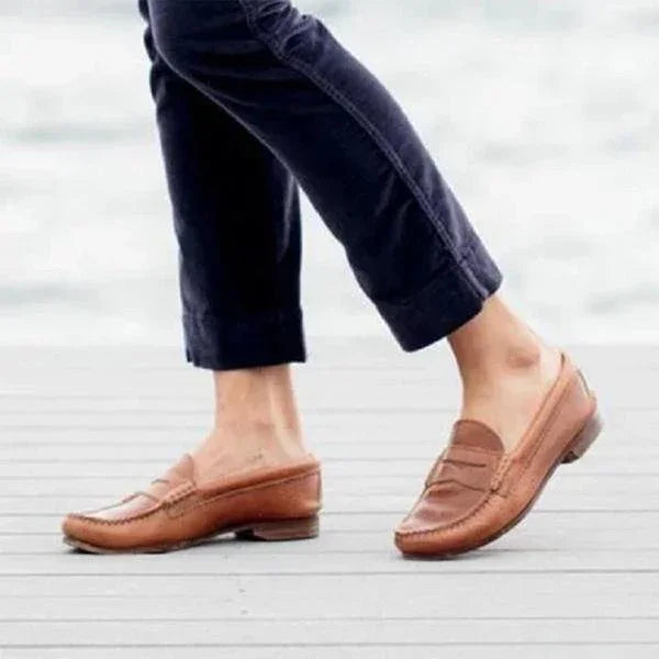 Person wearing brown leather slip-on loafers with navy cropped pants on wooden floor