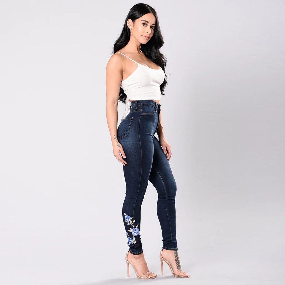 Woman in white crop top and dark blue embroidered skinny jeans with clear high heels on a plain background