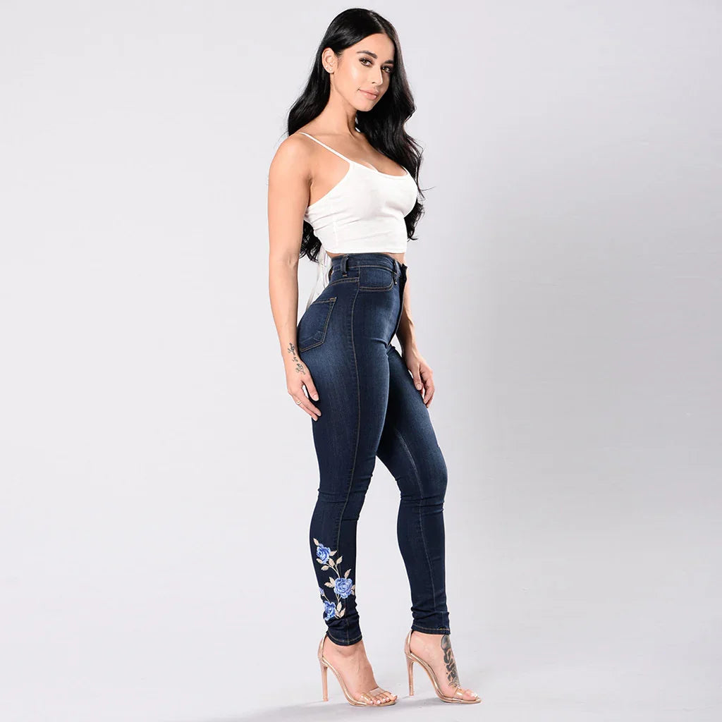 Woman in white crop top and dark blue embroidered skinny jeans with clear high heels on a plain background