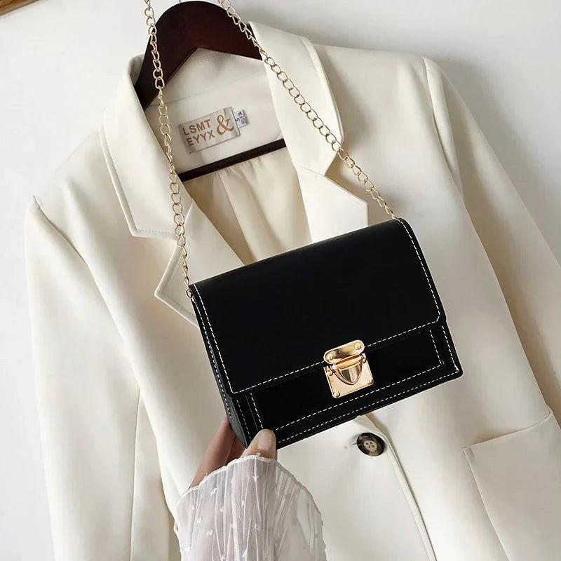 Black leather crossbody bag with gold clasp and chain strap held over white blazer on hanger