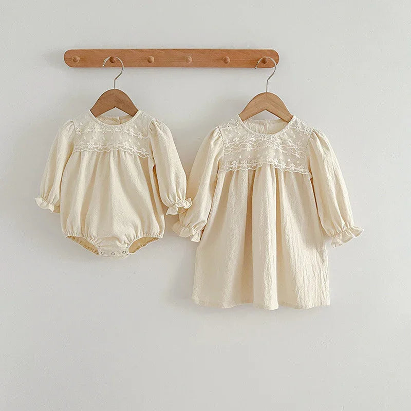 Two cream-colored baby dresses with floral embroidery, long sleeves, hanging on wooden hangers