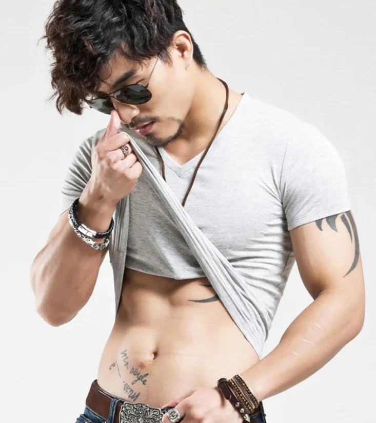 Muscular man with tattoos wearing sunglasses and bracelets, lifting gray t-shirt to show abs