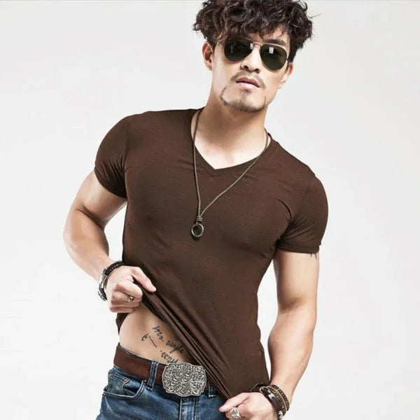 Man wearing brown fitted V-neck T-shirt, sunglasses, necklace, and jeans with ornate belt buckle, showing tattoo