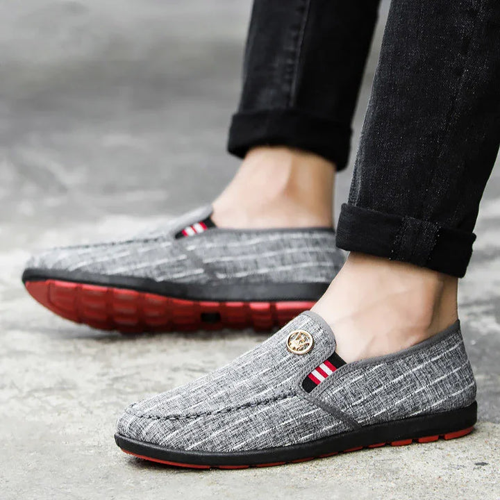 Casual gray slip-on men's shoes with red-striped detail and red soles worn with black rolled-up jeans