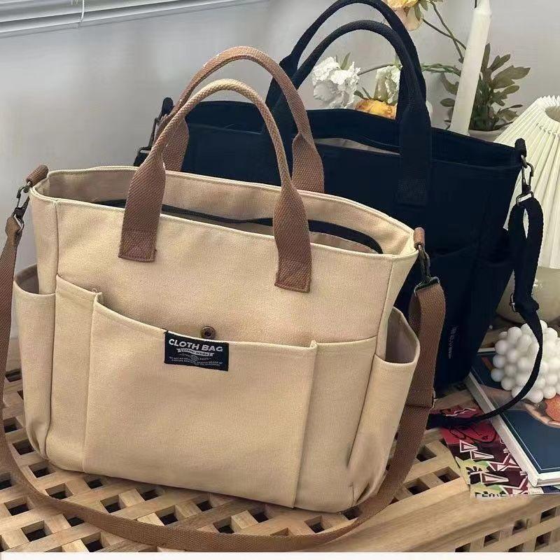 Beige and black canvas cloth bags with multiple pockets and handles on a wooden surface with flowers and decor