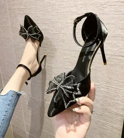 black high-heeled pointed-toe shoes with ankle straps and rhinestone bow detail worn with blue jeans