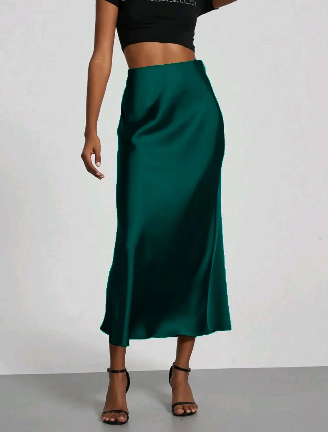 Woman wearing a teal satin midi skirt, black crop top, and black ankle strap heels on plain background