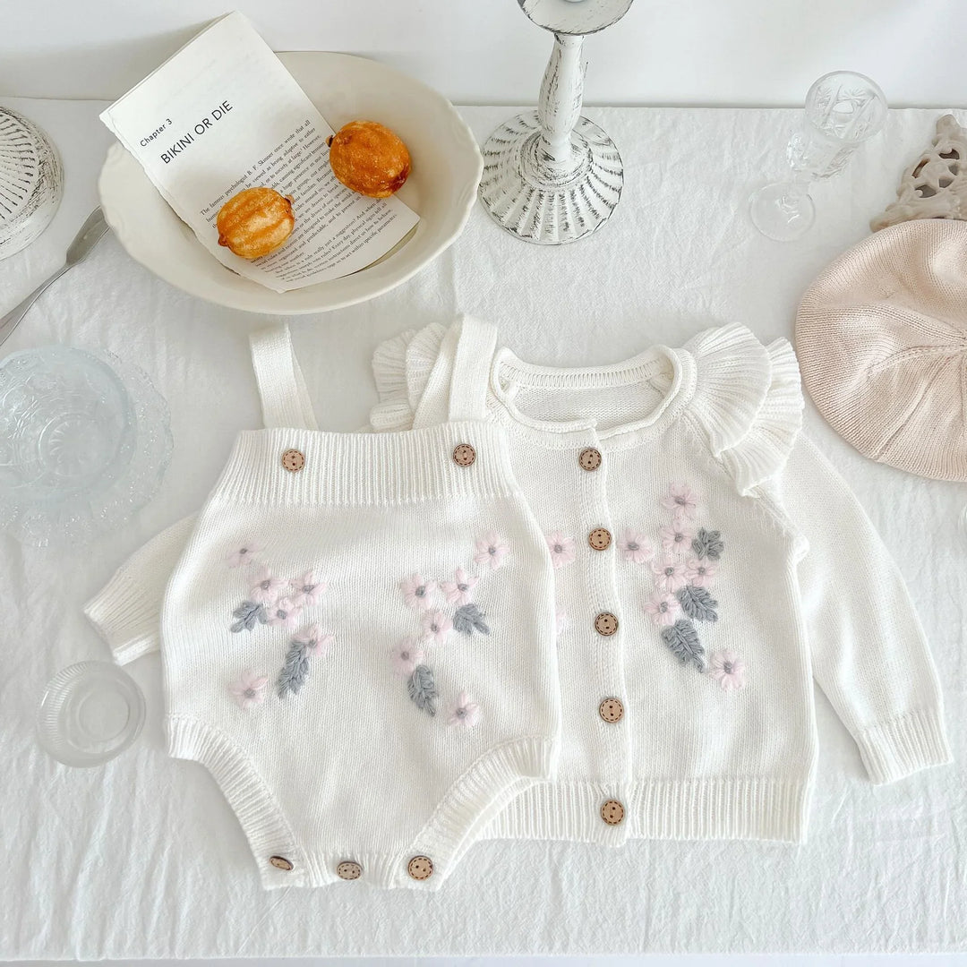 White knitted baby romper and cardigan with pink flowers and buttons on white cloth with beige hat