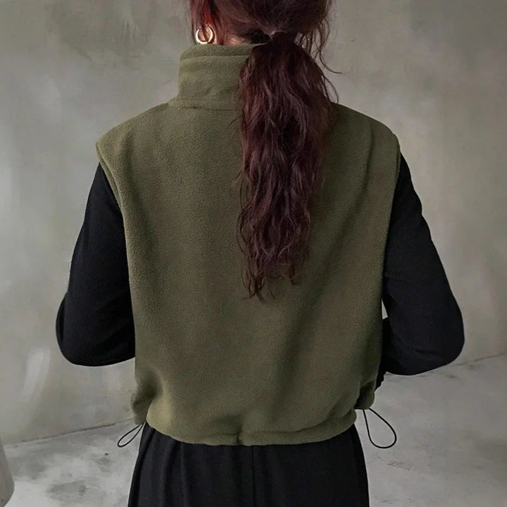 Woman with long brown hair wearing olive green fleece vest and black long-sleeve top indoors
