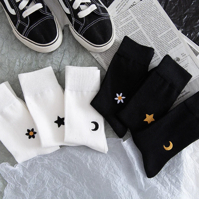 Black and white ankle socks with embroidered moon, star, and flower designs next to black sneakers and newspaper