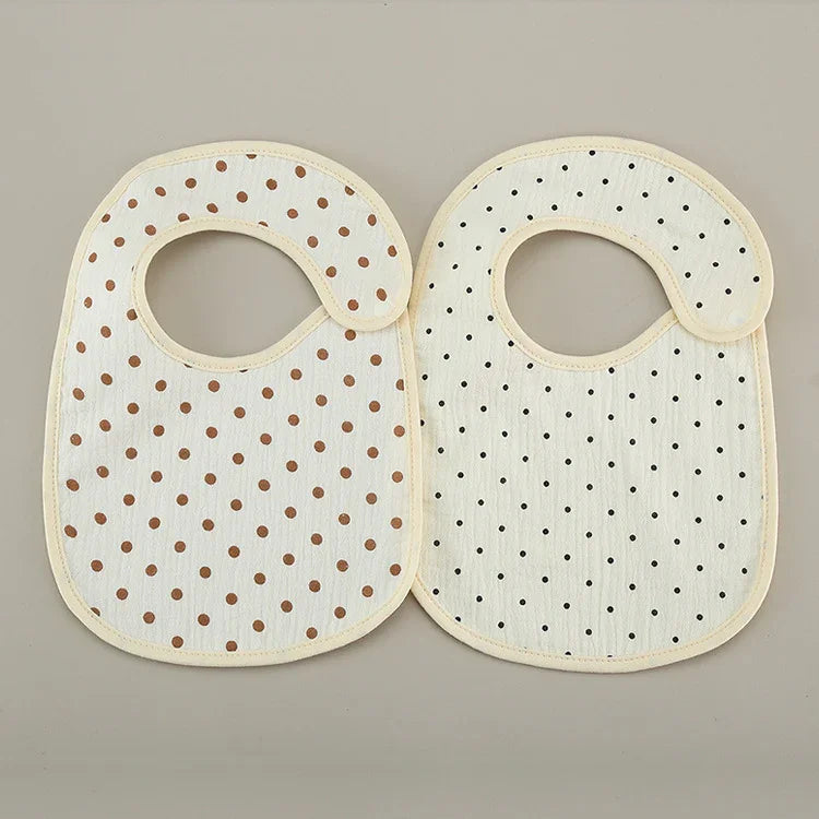 Two soft cotton baby bibs with beige trim, one with brown polka dots and one with black polka dots on a light background