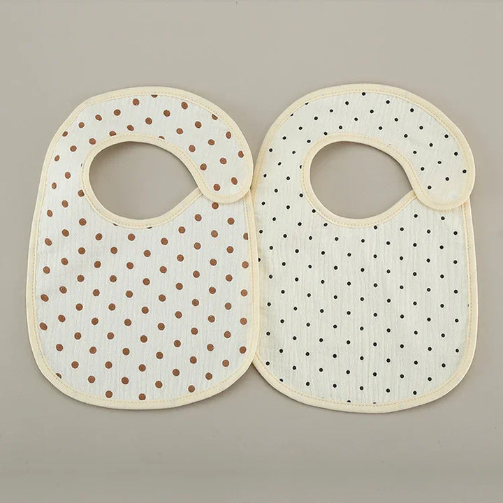 Two soft cotton baby bibs with beige trim, one with brown polka dots and one with black polka dots on a light background