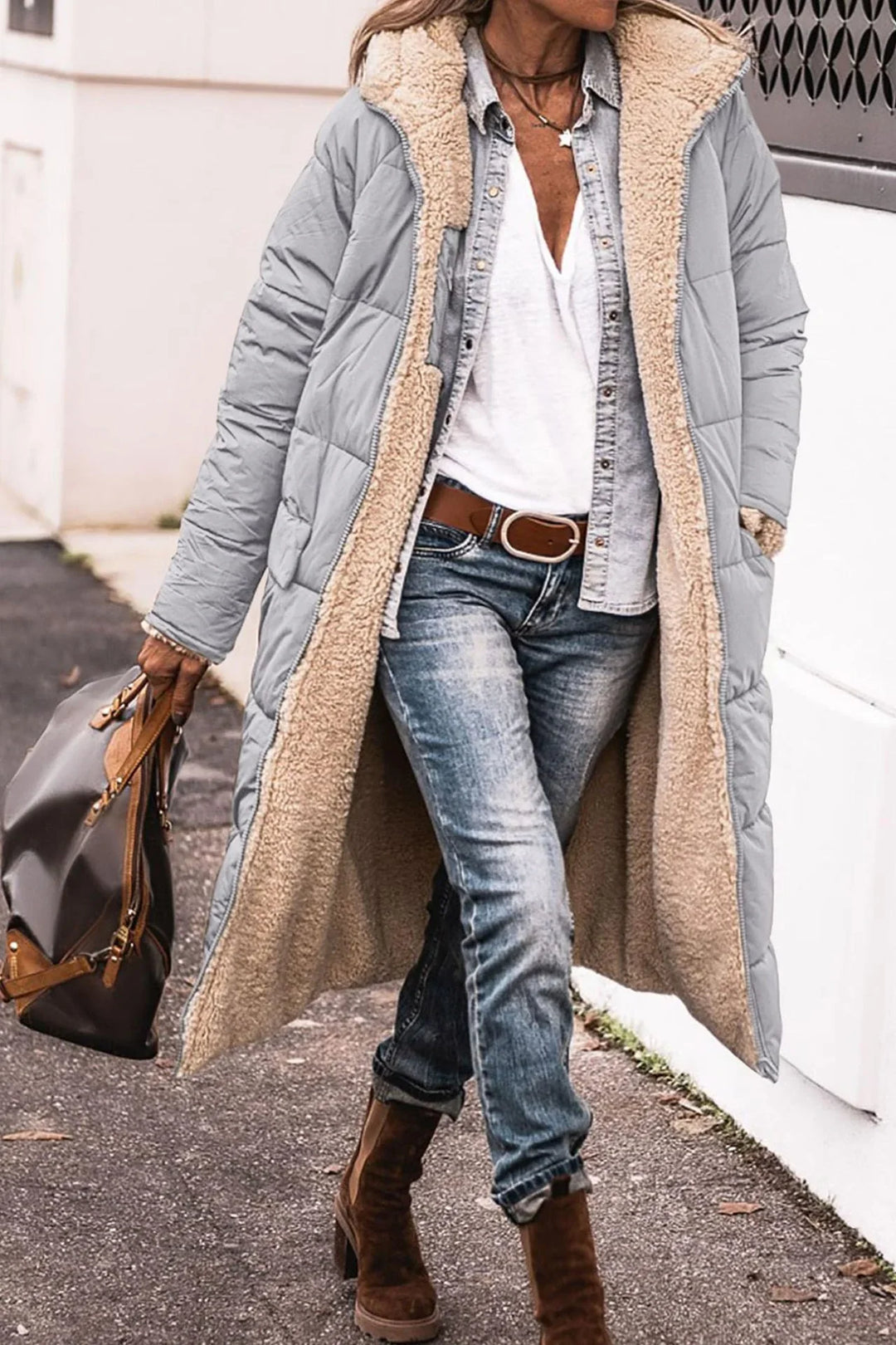 Woman wearing long gray quilted coat with beige sherpa lining, denim jeans, brown boots, and carrying brown handbag