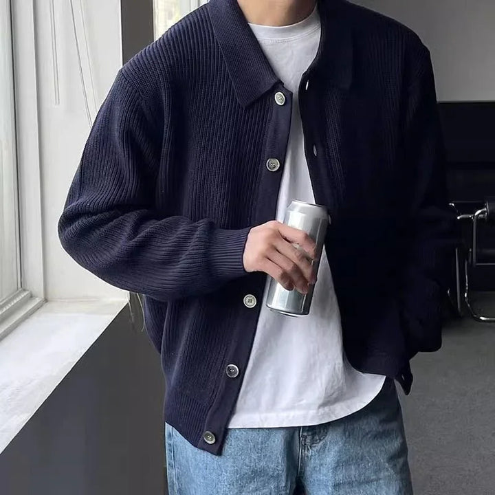 Man wearing navy blue ribbed cardigan over white t-shirt with jeans holding a silver can indoors