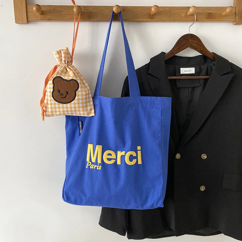 Blue tote bag with Merci Paris text, yellow checkered drawstring bag with bear patch, and black blazer on wooden hooks