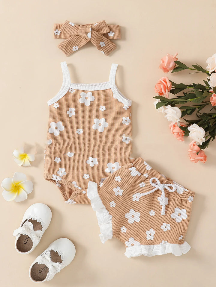 Beige baby outfit set with white flower print, sleeveless bodysuit, shorts with ruffles, matching headband, white baby shoes, and decorative flowers
