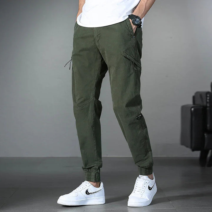Man wearing olive green cargo jogger pants with cuffs and white Nike sneakers on grey background